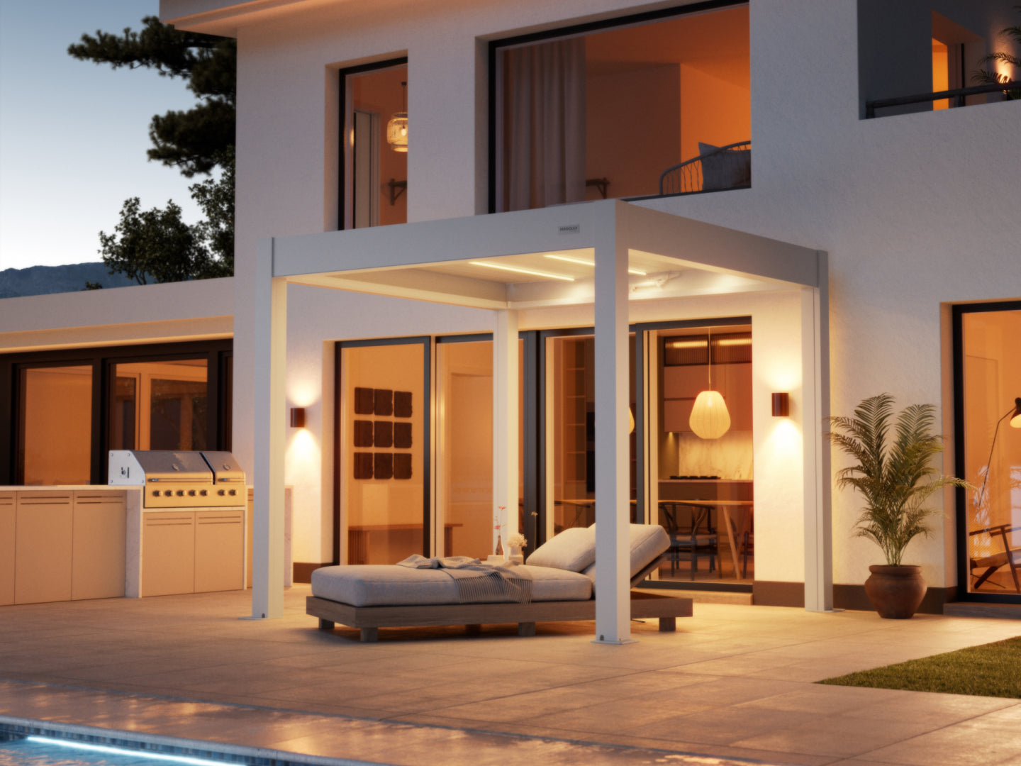  A contemporary white pergola with integrated LED strip lighting provides shade over a paved patio area next to a modern white stucco house at dusk. Underneath the pergola, a comfortable-looking chaise lounge with gray cushions is positioned for relaxation. The patio also features a built-in outdoor kitchen and grill on the left and a potted plant on the right. Large glass doors and windows reveal a warm, inviting interior with pendant lighting. A swimming pool's edge is visible in the foreground.