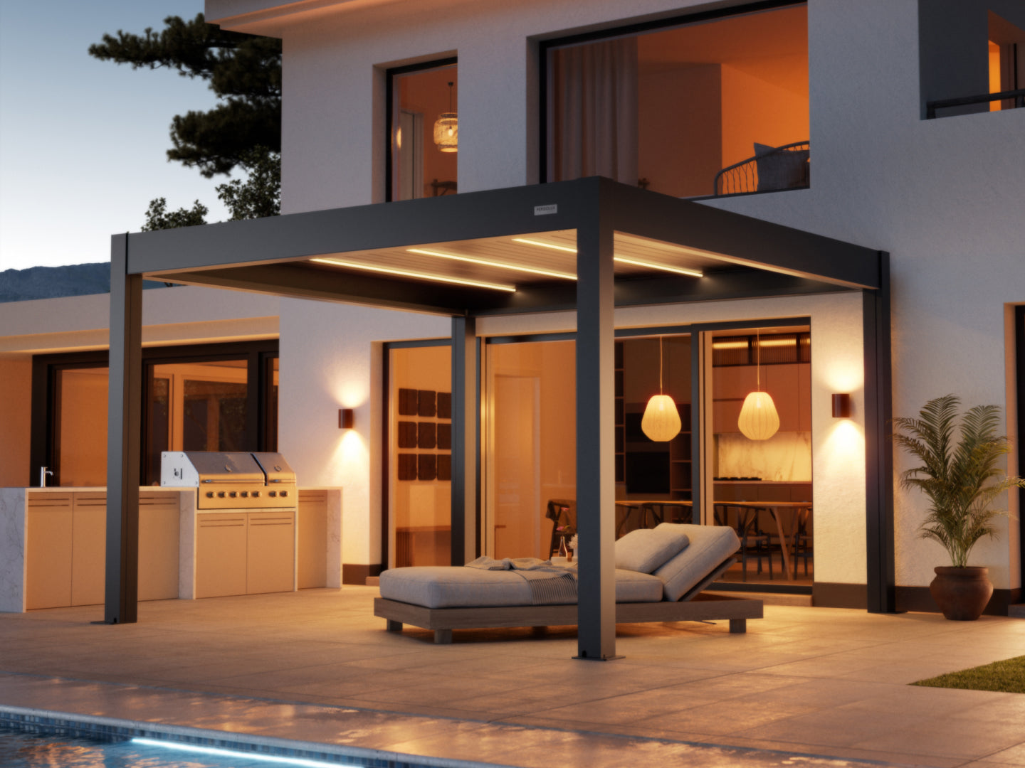 A dark gray, modern pergola with an adjustable louvered roof and integrated LED strip lighting covers a paved patio next to a contemporary white house at dusk. Beneath the pergola, a comfortable-looking chaise lounge with gray cushions is positioned. To the left, there is a built-in outdoor kitchen and grill area. The house has large glass doors and windows revealing a warm, inviting interior, which features decorative pendant lights. Accent wall sconces flank the glass doors, and a potted plant is visible 