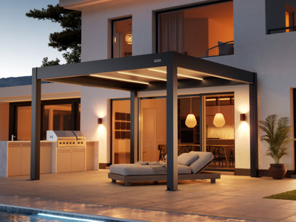 A dark gray, modern pergola with an adjustable louvered roof and integrated LED strip lighting covers a paved patio next to a contemporary white house at dusk. Beneath the pergola, a comfortable-looking chaise lounge with gray cushions is positioned. To the left, there is a built-in outdoor kitchen and grill area. The house has large glass doors and windows revealing a warm, inviting interior, which features decorative pendant lights. Accent wall sconces flank the glass doors, and a potted plant is visible 
