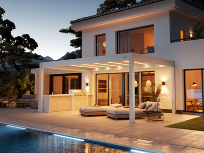 A sleek, modern, white pergola with integrated LED strip lighting provides shade over a patio area next to a contemporary white house. Beneath the pergola is a comfortable-looking gray outdoor chaise lounge. The house features large glass doors and windows that showcase warm, inviting interior lighting. On the patio, there are also a built-in outdoor grill area and a potted plant. The overall atmosphere is luxurious and serene, illuminated by the soft interior and accent lighting at dus